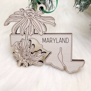 May include: A light-coloured wooden ornament shaped like the state of Maryland, featuring an engraved floral design and the word MARYLAND in black text. The ornament includes a green ribbon loop for hanging, showcasing detailed line art of flowers.
