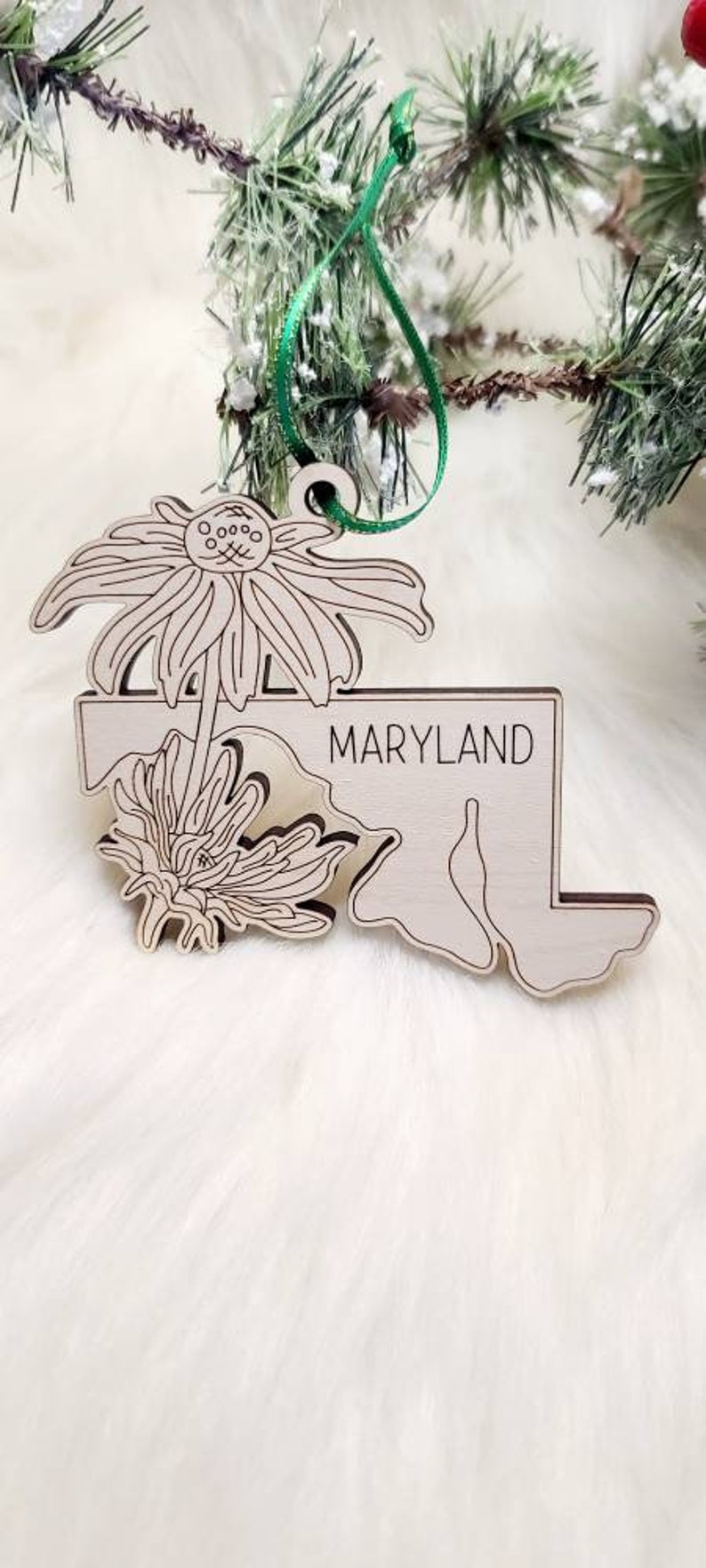 May include: A light-coloured wooden ornament shaped like the state of Maryland, featuring an engraved floral design and the word MARYLAND in black text. The ornament includes a green ribbon loop for hanging, showcasing detailed line art of flowers.