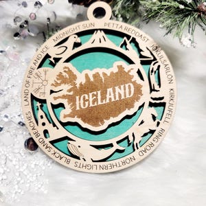 May include: A circular wooden ornament featuring a cutout map of Iceland with the text ICELAND in the center. The border includes engraved text and icons representing landmarks like a church, whale, and mountain, set against a teal background.