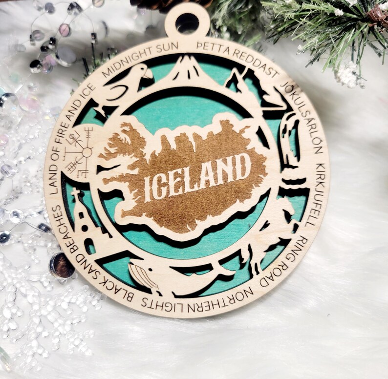 May include: A circular wooden ornament featuring a cutout map of Iceland with the text ICELAND in the center. The border includes engraved text and icons representing landmarks like a church, whale, and mountain, set against a teal background.
