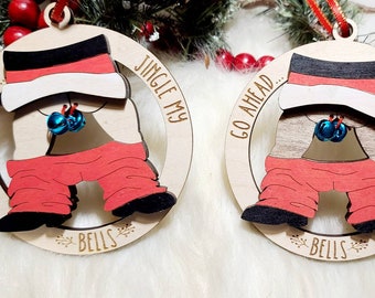 Puede incluir: Dos adornos circulares de madera con un diseño cortado a láser de piernas de Santa Claus con pantalones rojos y botas negras. El texto "GO AHEAD... JINGLE MY BELLS" está grabado en el marco. Cada adorno incluye dos pequeños cascabeles azules.