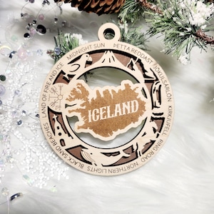 May include: A circular wooden ornament featuring a cutout map of Iceland in the center with the word ICELAND engraved. The border includes icons of a puffin, volcano, church, and whale, with text reading LAND OF FIRE AND ICE, MIDNIGHT SUN, PETTA REDDAST, JOKULSARLON, KIRJUFELL, RING ROAD, NORTHERN LIGHTS, and BLACK SAND BEACHES.