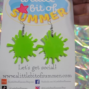 May include: A pair of lime green splatter paint earrings with silver hooks. The earrings are on a white card with the text "a little bit of summer" and social media information.
