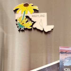 May include: A wooden magnet shaped like the state of Maryland, featuring the word MARYLAND in black text. The design includes two yellow black-eyed Susan flowers with green stems and leaves layered over the left side of the state outline.