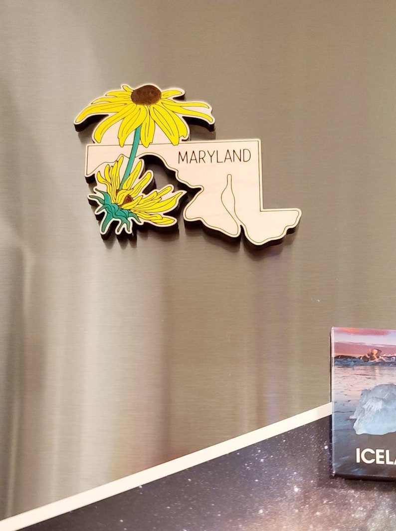 May include: A wooden magnet shaped like the state of Maryland, featuring the word MARYLAND in black text. The design includes two yellow black-eyed Susan flowers with green stems and leaves layered over the left side of the state outline.
