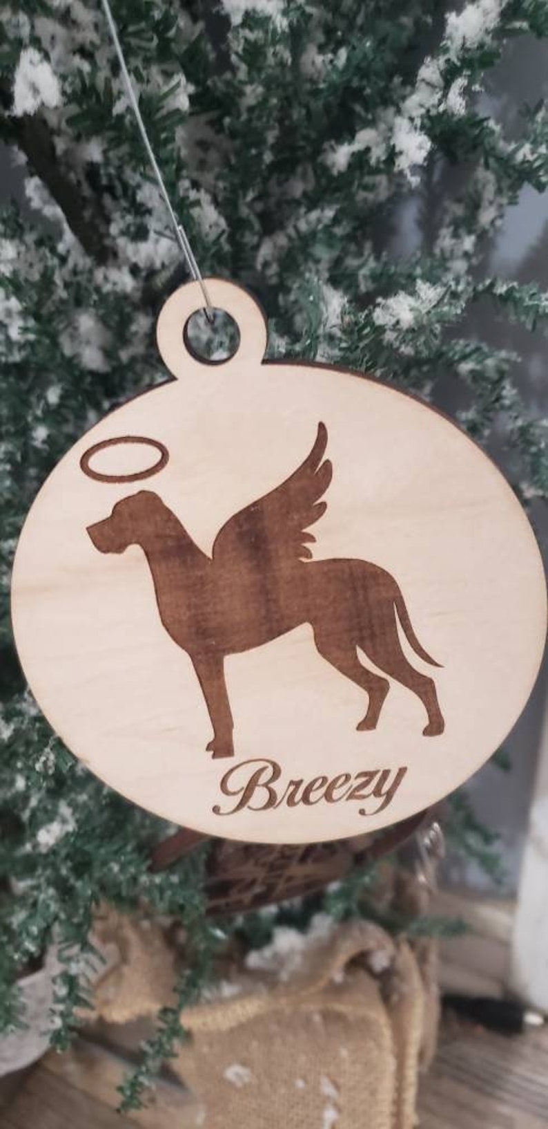 K&ouml;nnte beinhalten: Ein Holz-Anh&auml;nger mit einer Hundesilhouette mit Engelsfl&uuml;geln und einem Heiligenschein. Der Hund ist braun und der Anh&auml;nger ist hellbraun. Der Anh&auml;nger tr&auml;gt den Namen "Breezy" eingraviert.