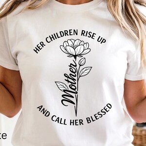 Puede incluir: Camiseta blanca con un diseño floral y las palabras en negro "HER CHILDREN RISE UP AND CALL HER BLESSED". La palabra "Mother" está escrita verticalmente en cursiva. La palabra "White" está escrita en la esquina inferior izquierda.