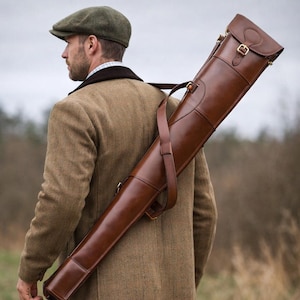 May include: A brown leather gun case with a shoulder strap, carried over the shoulder. The case has a flap closure with a brass buckle and is made of stitched leather. The case is long and slender, designed to hold a long gun.