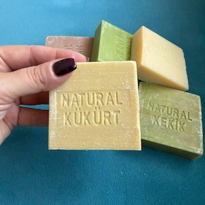 Könnte beinhalten: Mehrere Seifenstücke aus Naturseife in verschiedenen Grün-, Beige- und Brauntönen liegen auf einer blauen Oberfläche. Ein Seifenstück, das von einer Hand gehalten wird, ist mit den Worten "NATURAL KÜKÜRT" versehen. Auch andere Seifenstücke sind mit Text versehen.