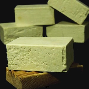 Puede incluir: Varias barras de jabón rectangulares de color verde claro con una superficie texturizada. Las barras están apiladas, una de ellas sobre un jabonera de madera. El aspecto natural y artesanal sugiere ingredientes naturales.