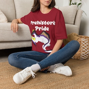 Puede incluir: Una camiseta de manga corta de color rojo oscuro con el texto blanco "Prehistoric Pride" sobre un gráfico de un dinosaurio blanco, morado y amarillo con una vela moteada en la espalda, sosteniendo una bandera con los mismos colores morado, blanco y amarillo.