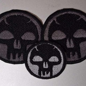 May include: Three circular embroidered patches featuring a stylised black skull design on a dark grey background. The two larger patches are positioned at the top, while a smaller patch is centred below them, all with black stitched borders.