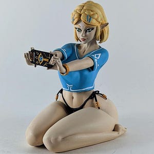 May include: A figurine of a character with blonde hair and pointed ears, wearing a blue top and black bikini bottoms, holding a black device with a gold design. The figure is kneeling on a white surface.