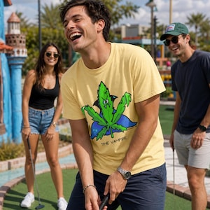 May include: A pale yellow short-sleeve t-shirt featuring a graphic of a green leaf character with vampire fangs and a blue cape, with the text "TIME VAMPIRE" printed in black below the illustration on the front of the garment.