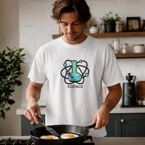 May include: A white short-sleeved t-shirt featuring a graphic of a light blue laboratory flask centered within a black atom symbol. Below the graphic, the word SCIENCE is printed in bold black capital letters. The shirt has a crew neckline.