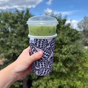 Puede incluir: Una taza de plástico transparente llena de té helado verde, cubierta con una tapa transparente, sostenida en una funda de taza con estampado en blanco y negro. La funda tiene un diseño repetitivo. El fondo es un cielo azul brillante y árboles verdes.