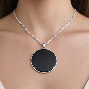 Puede incluir: Un collar de cadena plateada con un gran colgante redondo negro. El colgante tiene un acabado pulido y brillante y está engastado en un bisel plateado. El collar se muestra en el cuello.