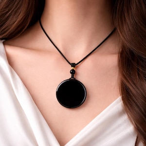 Puede incluir: Un collar con colgante negro con una piedra pulida y redonda. El colgante cuelga de un cordón negro con una pequeña cuenta dorada. El collar se muestra sobre una camisa blanca.