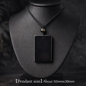 May include: A black rectangular pendant necklace displayed on a black mannequin. The pendant is approximately 2 inches by 1.2 inches. The necklace has a black cord and a decorative bead near the clasp. The background is dark.