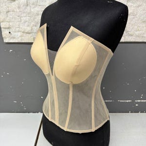 May include: A beige corset with a deep V-neckline and sheer mesh panels. The corset features padded cups and vertical boning for structure. It is displayed on a black mannequin against a neutral background.