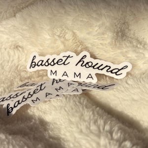 May include: White stickers with black cursive text that reads "basset hound MAMA". Several stickers are scattered on a soft, cream-colored surface. The stickers are in a script font.