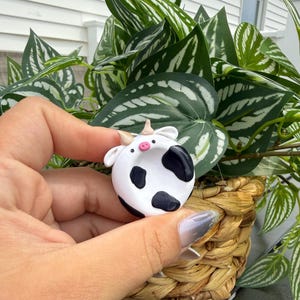 May include: A small, circular, white clay charm featuring a black cow-spotted pattern, two small tan horns, and a tiny pink snout. The charm has a smooth, rounded surface with two black dot eyes positioned above the pink nose, creating a simple animal face.