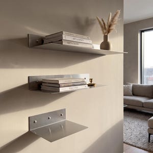Puede incluir: Tres estantes de pared de metal cepillado con una superficie horizontal plana y una placa trasera vertical asegurada con tornillos visibles. El estante superior sostiene tres libros y un jarrón beige con hierba seca. El estante central sostiene tres libros y una vela.
