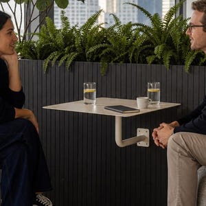 Op de afbeelding: Een aan de muur gemonteerde, vierkante tafel van lichtbeige metaal met één steunarm. Op de tafel staan twee glazen water met schijfjes citroen en een klein espressokopje. De tafel is bevestigd aan een zwart wandpaneel met verticale latten.