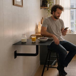 May include: A black wall-mounted folding table with a thin metal surface and a sturdy L-shaped support bracket. The table is positioned against a light-colored tiled wall, providing a compact dining space with a small glass bottle and bowl.