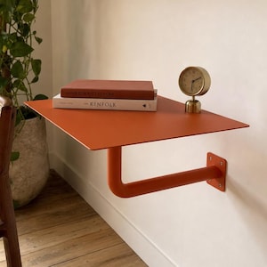Peut inclure: Une étagère murale flottante au fini orange brûlé mat, dotée d'une surface rectangulaire plate soutenue par un bras tubulaire unique fixé à une plaque de base carrée. Deux livres et une petite horloge analogique dorée sont posés dessus.