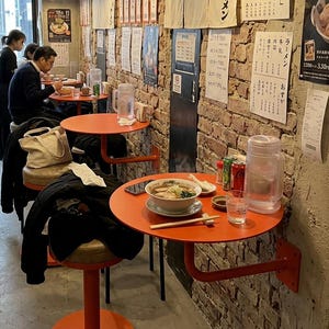 May include: A restaurant interior featuring round orange tables mounted to a brick wall, each paired with a matching orange stool. A bowl of ramen with chopsticks sits on the foreground table, alongside a clear water pitcher and a glass.