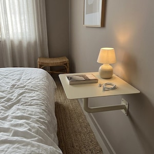 May include: A wall-mounted floating bedside shelf in a matte off-white finish, featuring a flat rectangular surface supported by a single L-shaped metal bracket. A small ceramic lamp, a closed book, and white wired earbuds rest on the shelf.