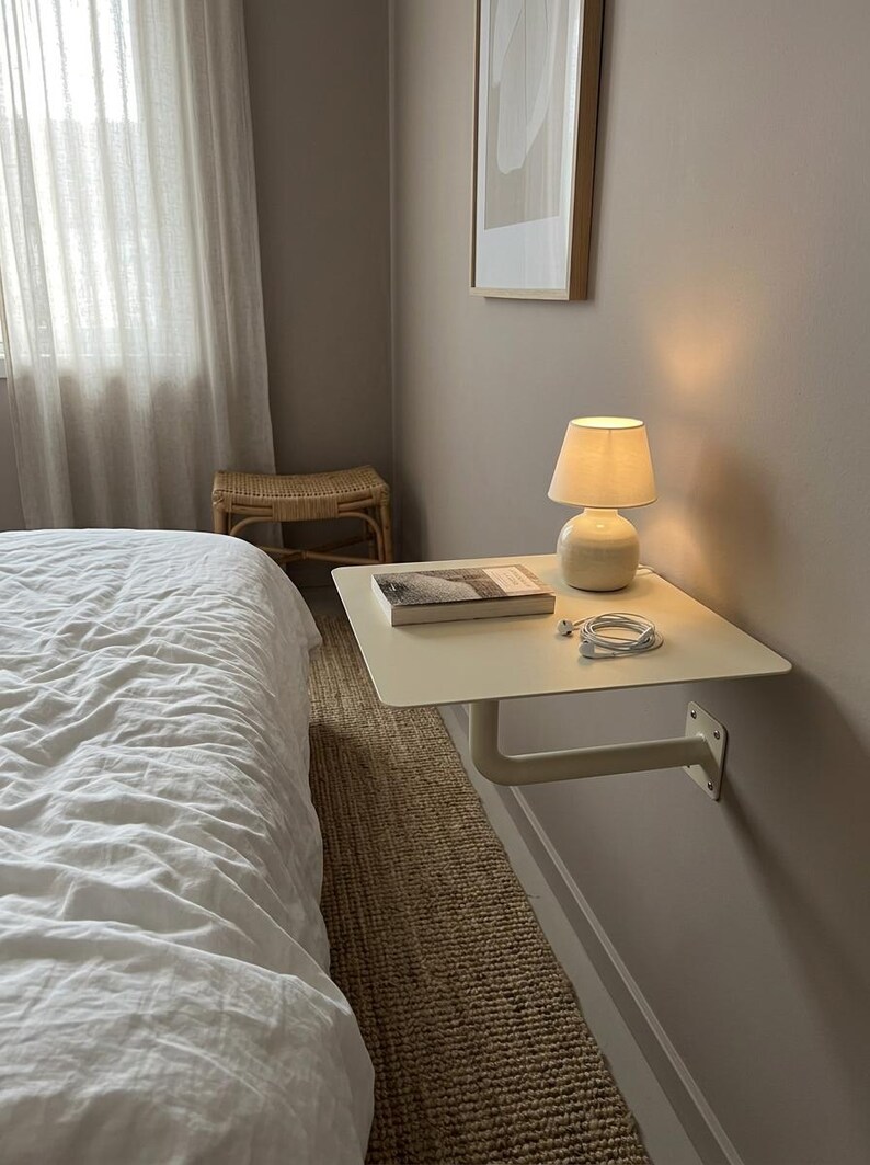 May include: A wall-mounted floating bedside shelf in a matte off-white finish, featuring a flat rectangular surface supported by a single L-shaped metal bracket. A small ceramic lamp, a closed book, and white wired earbuds rest on the shelf.