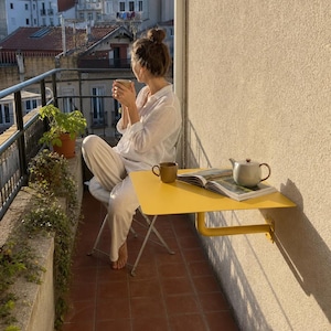 May include: A bright yellow wall-mounted balcony table with a smooth rectangular surface and a sturdy support bracket. On the table sits a ceramic teapot and an open book. The table is attached to a beige exterior wall on a tiled balcony.