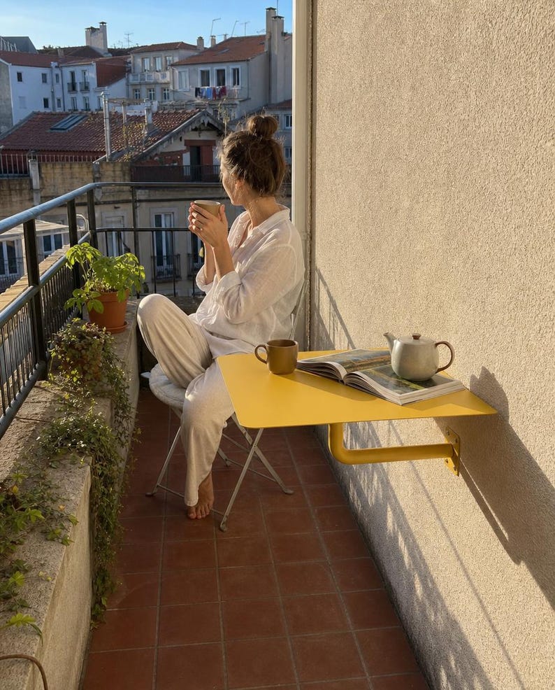 May include: A bright yellow wall-mounted balcony table with a smooth rectangular surface and a sturdy support bracket. On the table sits a ceramic teapot and an open book. The table is attached to a beige exterior wall on a tiled balcony.