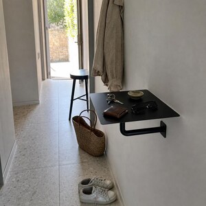May include: A matte black metal wall-mounted shelf featuring a flat rectangular surface supported by a single L-shaped bracket. The shelf holds a brown leather wallet, a set of metal keys, a small ceramic bowl, and black-framed eyeglasses.