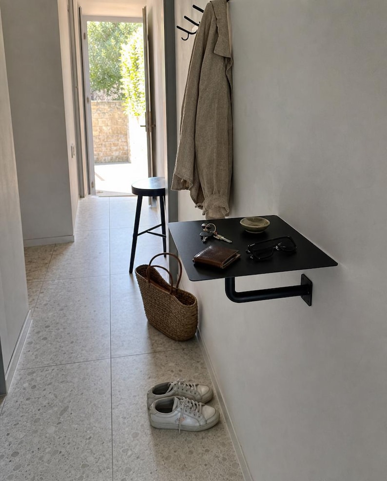 May include: A matte black metal wall-mounted shelf featuring a flat rectangular surface supported by a single L-shaped bracket. The shelf holds a brown leather wallet, a set of metal keys, a small ceramic bowl, and black-framed eyeglasses.