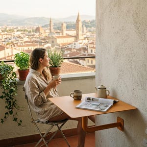 May include: A wall-mounted, square, orange metal balcony table with a single support arm, featuring a ceramic mug and a teapot on its surface, positioned on a balcony with a view of a city skyline in the background.