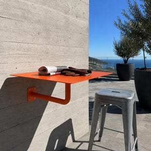 May include: A bright orange wall-mounted metal shelf with a single support arm attached to a concrete wall. A folded newspaper and a pair of brown leather gloves rest on the flat surface. A metallic gray stool stands on the ground nearby.