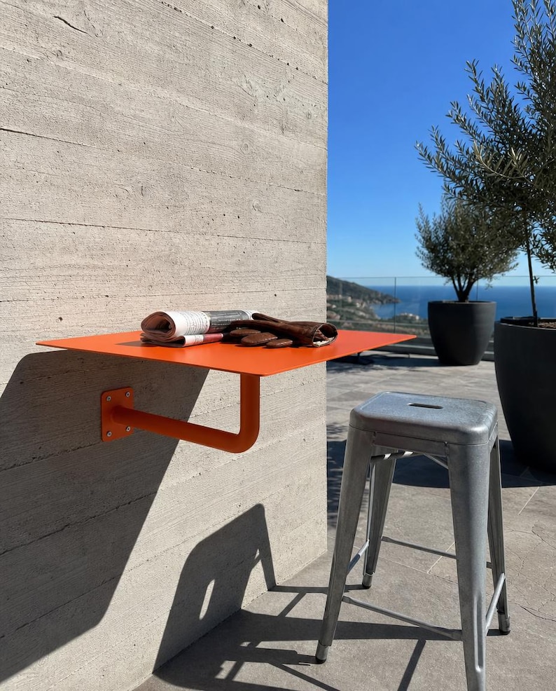 May include: A bright orange wall-mounted metal shelf with a single support arm attached to a concrete wall. A folded newspaper and a pair of brown leather gloves rest on the flat surface. A metallic gray stool stands on the ground nearby.