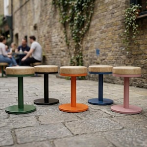 Può includere: Cinque sgabelli con sedute rotonde dalla texture color marrone chiaro e basi metalliche cilindriche in verde, nero, arancione, blu e rosa. Ogni sgabello presenta una base circolare piatta e una colonna di supporto centrale, allineati su una superficie in pietra.