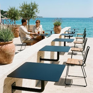 May include: A series of black metal square tables mounted to a stone wall, each paired with a black metal chair featuring light wood slats on the seat and backrest, arranged in a row along a terrace overlooking the blue sea.