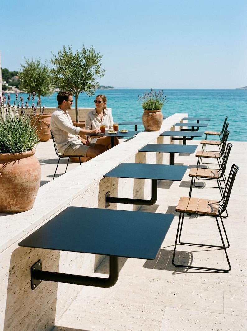May include: A series of black metal square tables mounted to a stone wall, each paired with a black metal chair featuring light wood slats on the seat and backrest, arranged in a row along a terrace overlooking the blue sea.