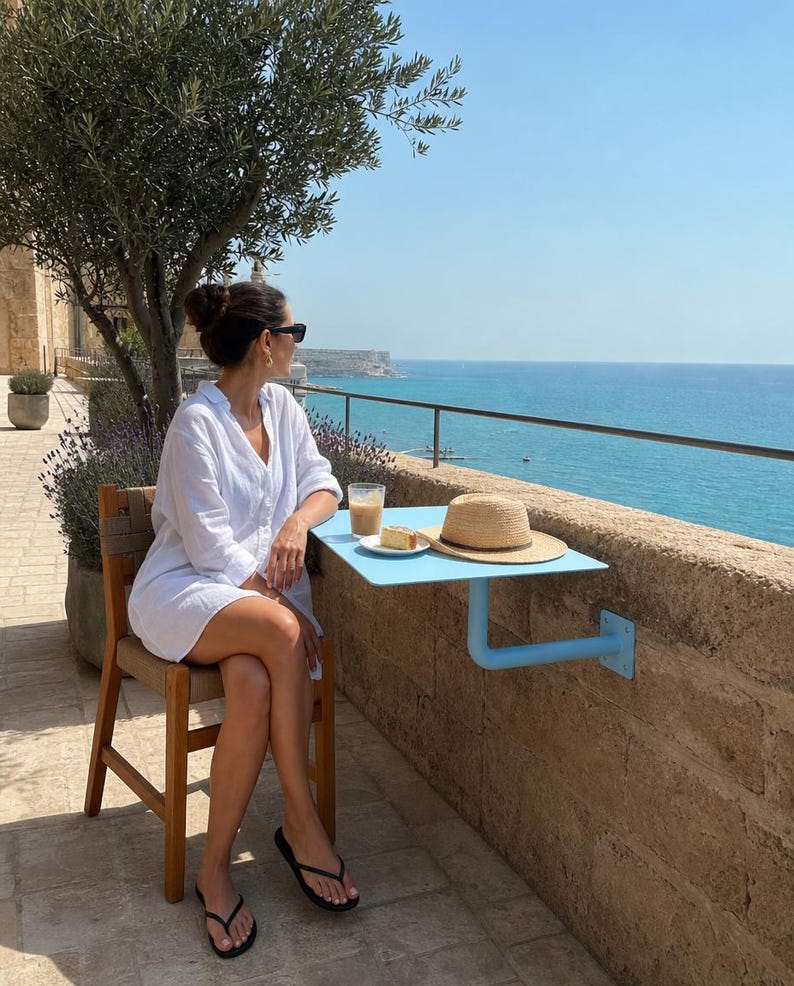May include: A light blue metal balcony table mounted to a stone wall, featuring a flat rectangular surface and a sturdy L-shaped support bracket. The table holds a glass of iced coffee, a small plate with a pastry, and a straw hat with a brown band.
