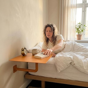 May include: A wall-mounted orange bedside shelf featuring a flat rectangular surface and a sturdy L-shaped support bracket attached directly to the wall. The shelf holds a small metallic alarm clock, a ceramic mug, and a book.