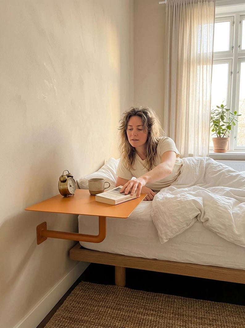 May include: A wall-mounted orange bedside shelf featuring a flat rectangular surface and a sturdy L-shaped support bracket attached directly to the wall. The shelf holds a small metallic alarm clock, a ceramic mug, and a book.