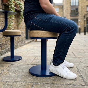 Puede incluir: Un taburete con un asiento circular de color tostado texturizado y una base de metal azul marino sólido que presenta un soporte cilíndrico central y una placa de suelo plana y redonda. Incluye un taburete a juego en el fondo.