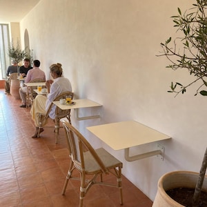 May include: A series of square, off-white metal tables mounted directly to a smooth beige wall, each paired with a woven rattan chair featuring a light-colored seat. The floor is covered in terracotta tiles, and a potted olive tree stands nearby.