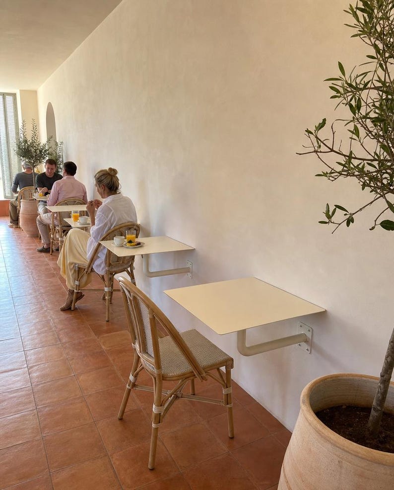 May include: A series of square, off-white metal tables mounted directly to a smooth beige wall, each paired with a woven rattan chair featuring a light-colored seat. The floor is covered in terracotta tiles, and a potted olive tree stands nearby.
