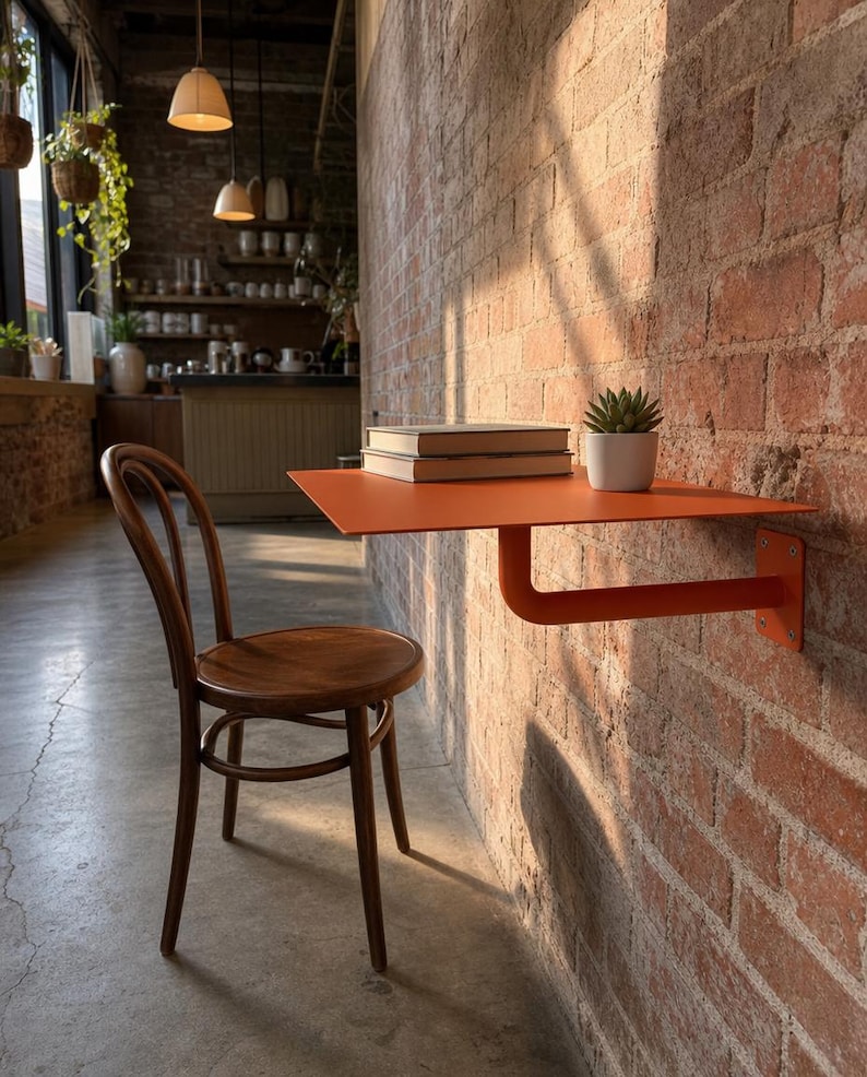 May include: A wall-mounted orange metal desk with a single support bracket attached to a brick wall. A dark brown wooden chair with a curved backrest is positioned in front of the desk. Two stacked books and a small potted succulent sit on top.